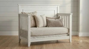 A photorealistic view of the farmhouse 4-in-1 crib converted into a daybed, showing the front slatted rail removed to create a sophisticated, open seating area while retaining the distressed white wood texture and solid headboard.